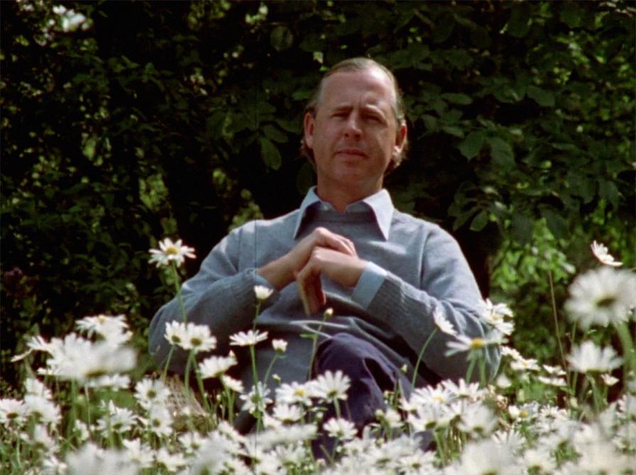 Klaus Rifbjerg siddende i blå skjorte i en have med hvide blomster.