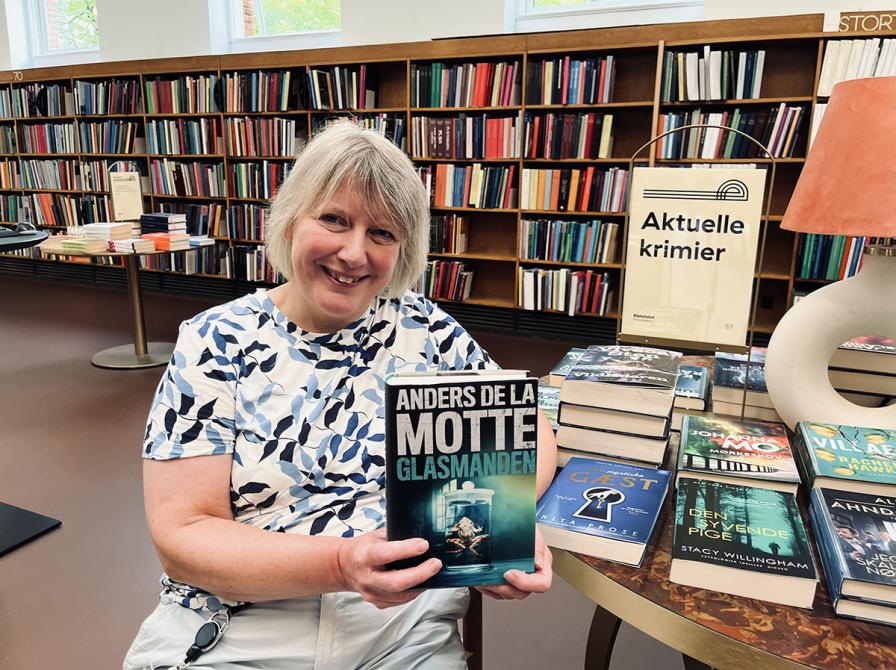 Kvindelig bibliotekar siddende med bogen Glasmanden ved siden af et bord med krimibøger.